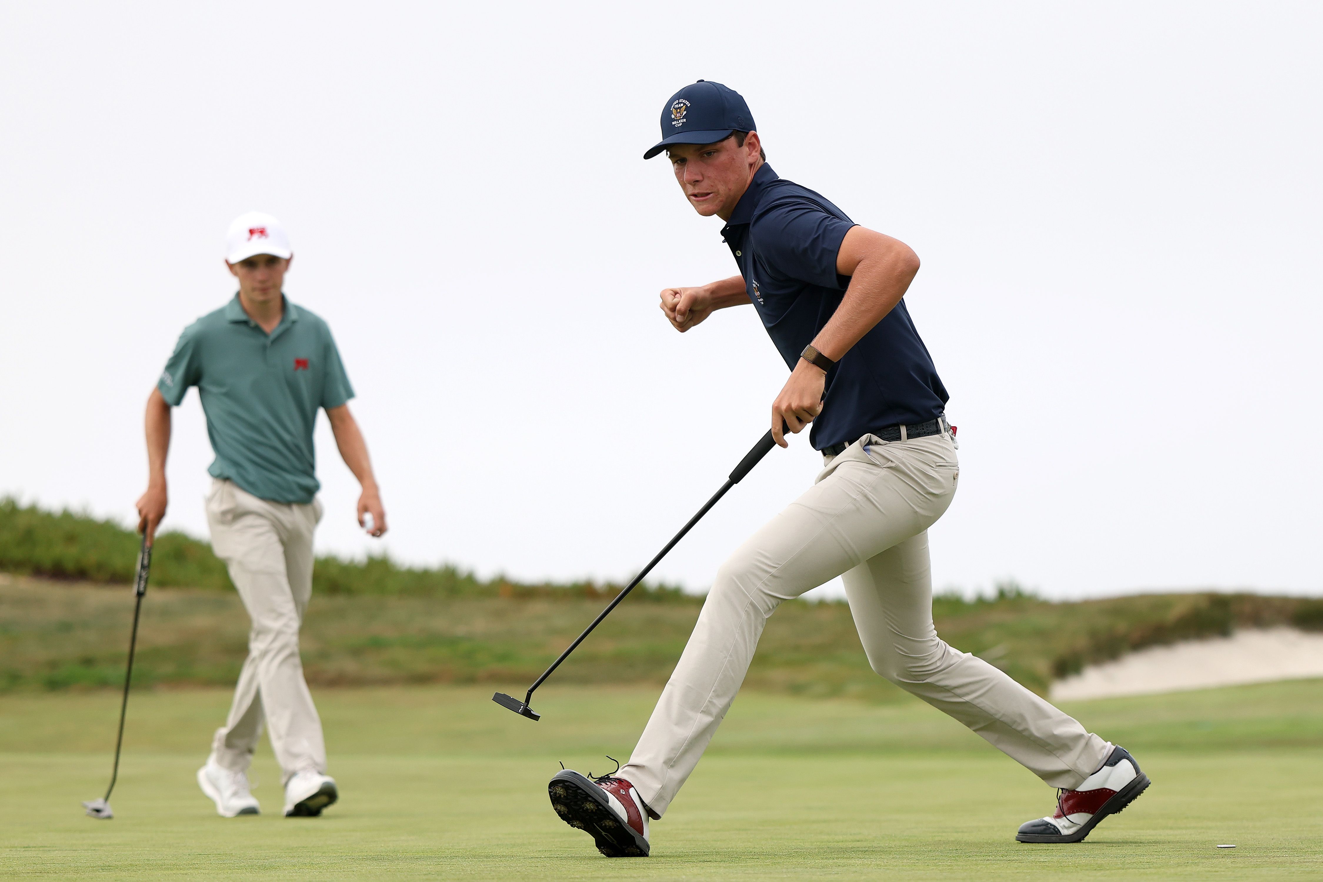 Golfer in blue shirt celebrates a successful putt on the green, watched by another player.