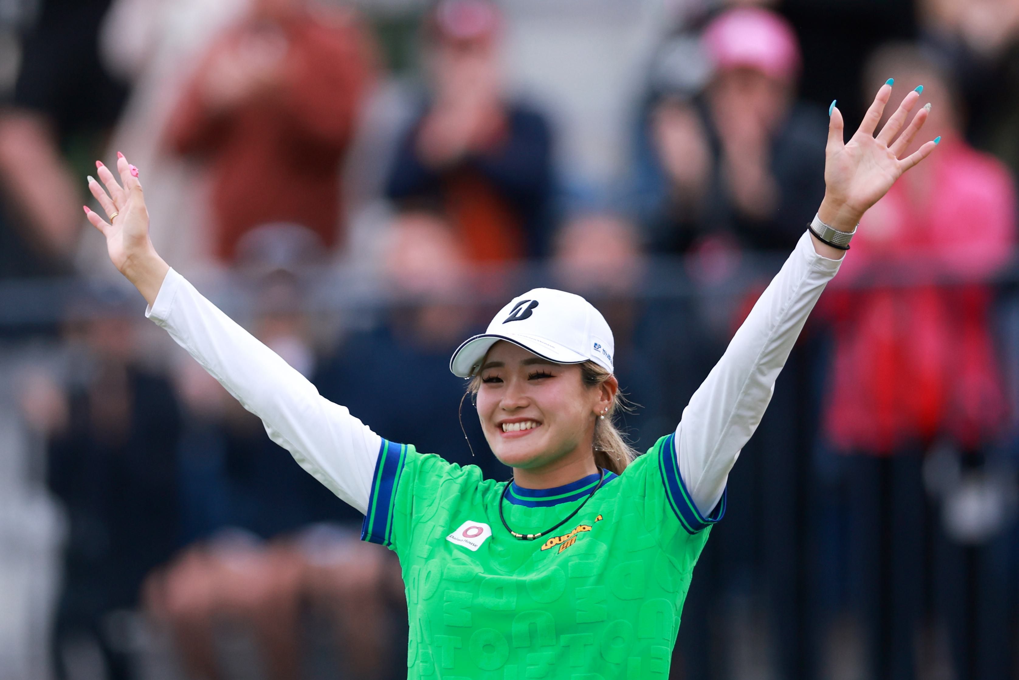 A woman athlete in a green shirt and white cap celebrates with arms raised.