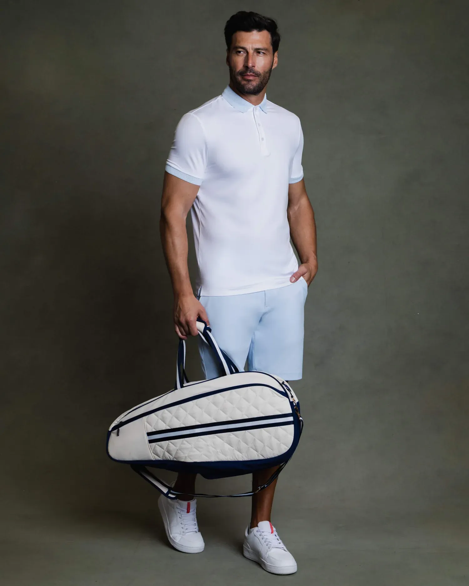 A stylish man in white athletic wear holds a tennis bag on a gray background.