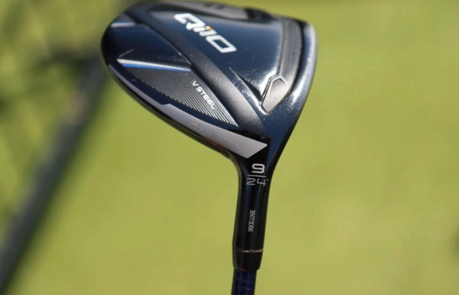 A dark golf club head with metallic accents against a blurred green background.