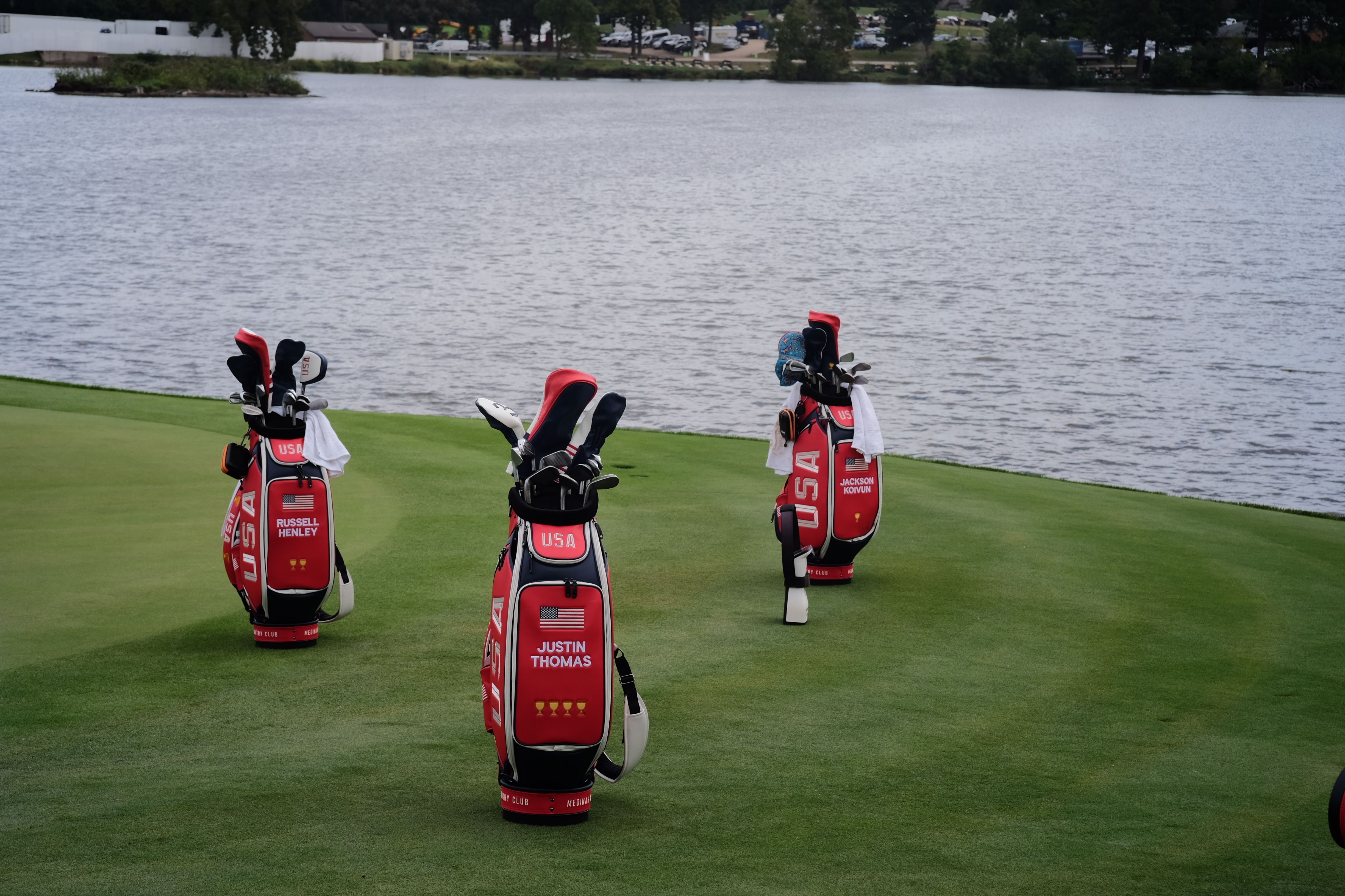 Three red and white golf bags stand on a green course next to a calm lake.