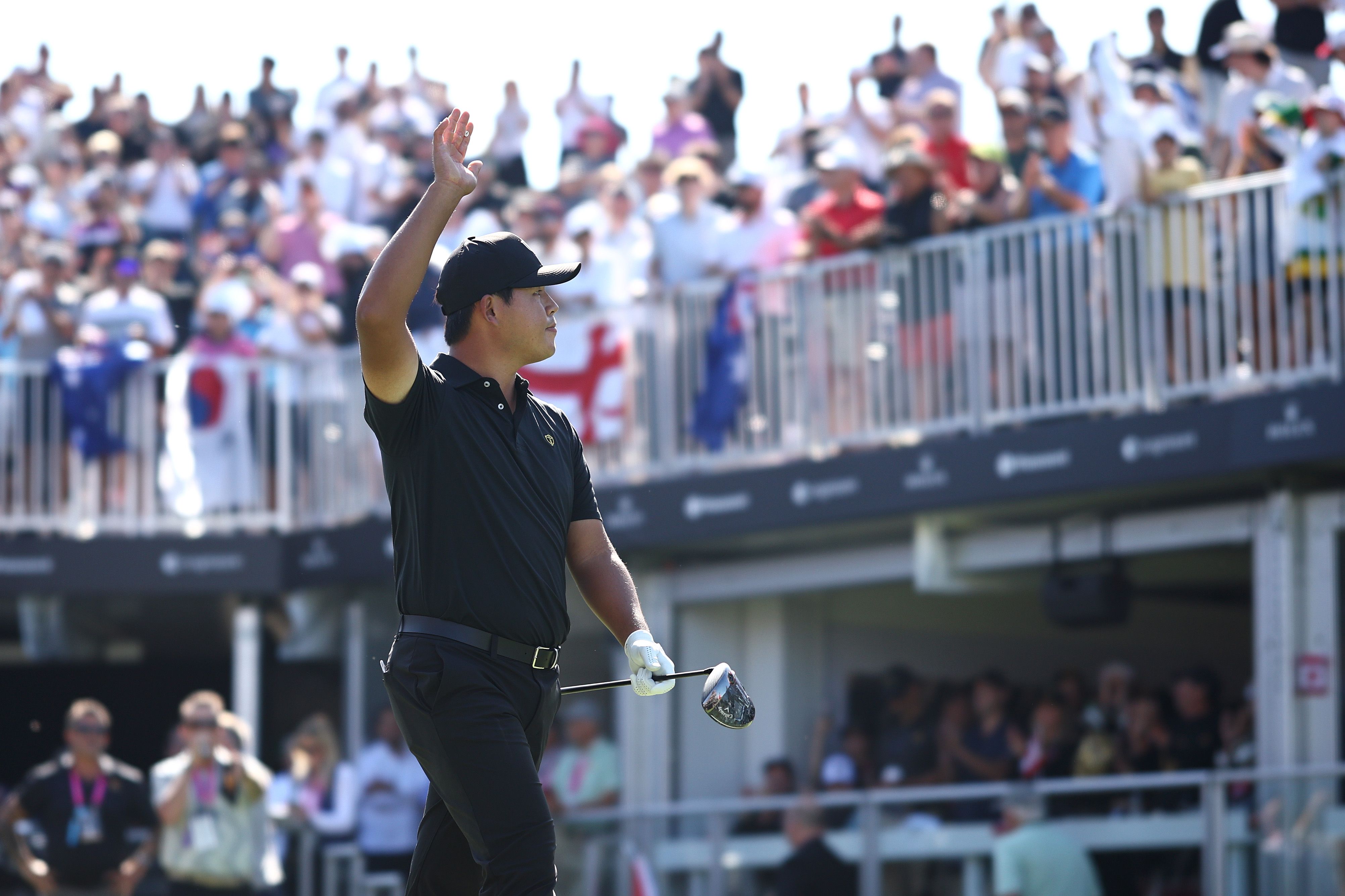 Golfer in black cap and shirt waves to cheering spectators from the course.