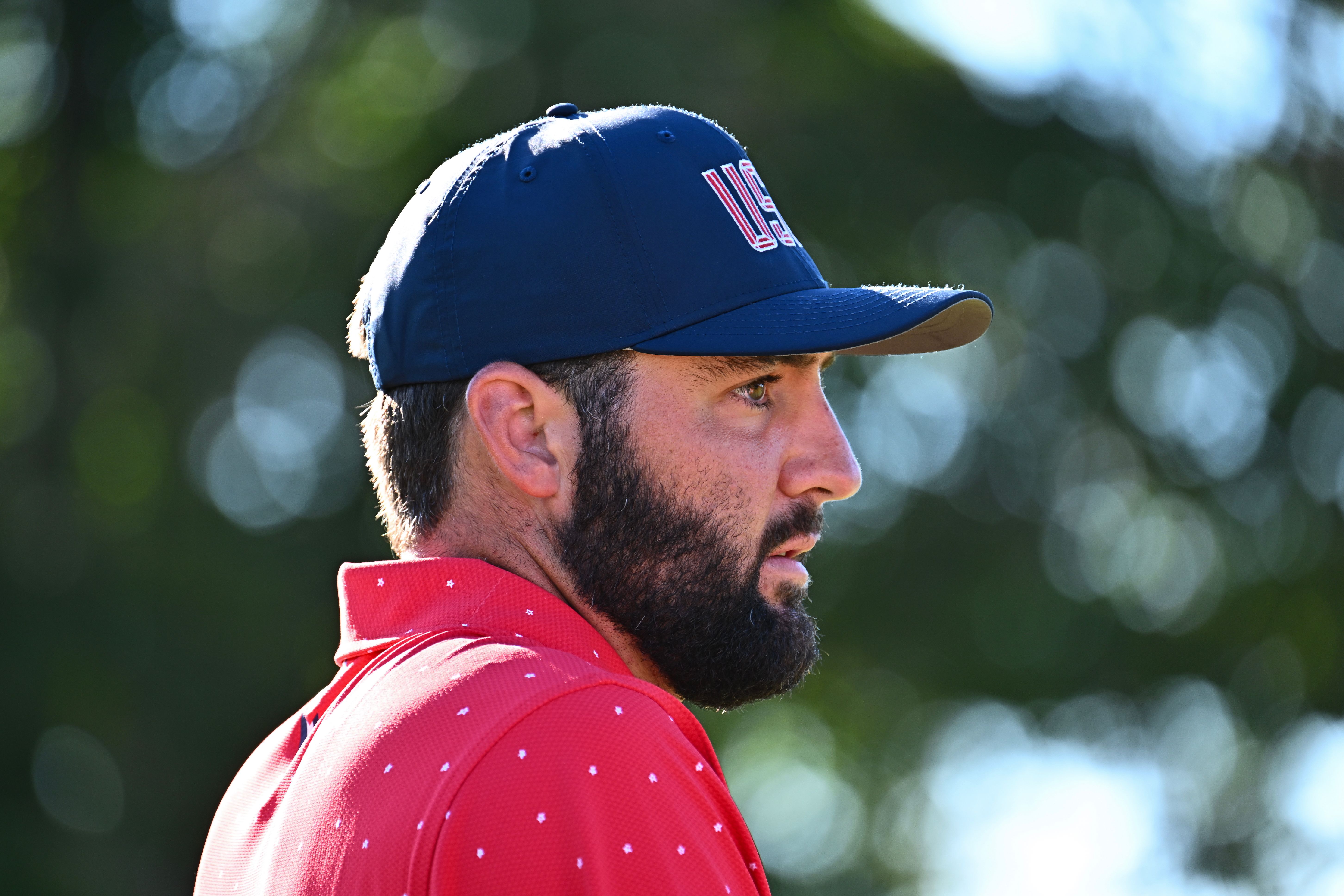 A bearded man wearing a red shirt and navy "USA" baseball cap looks right.