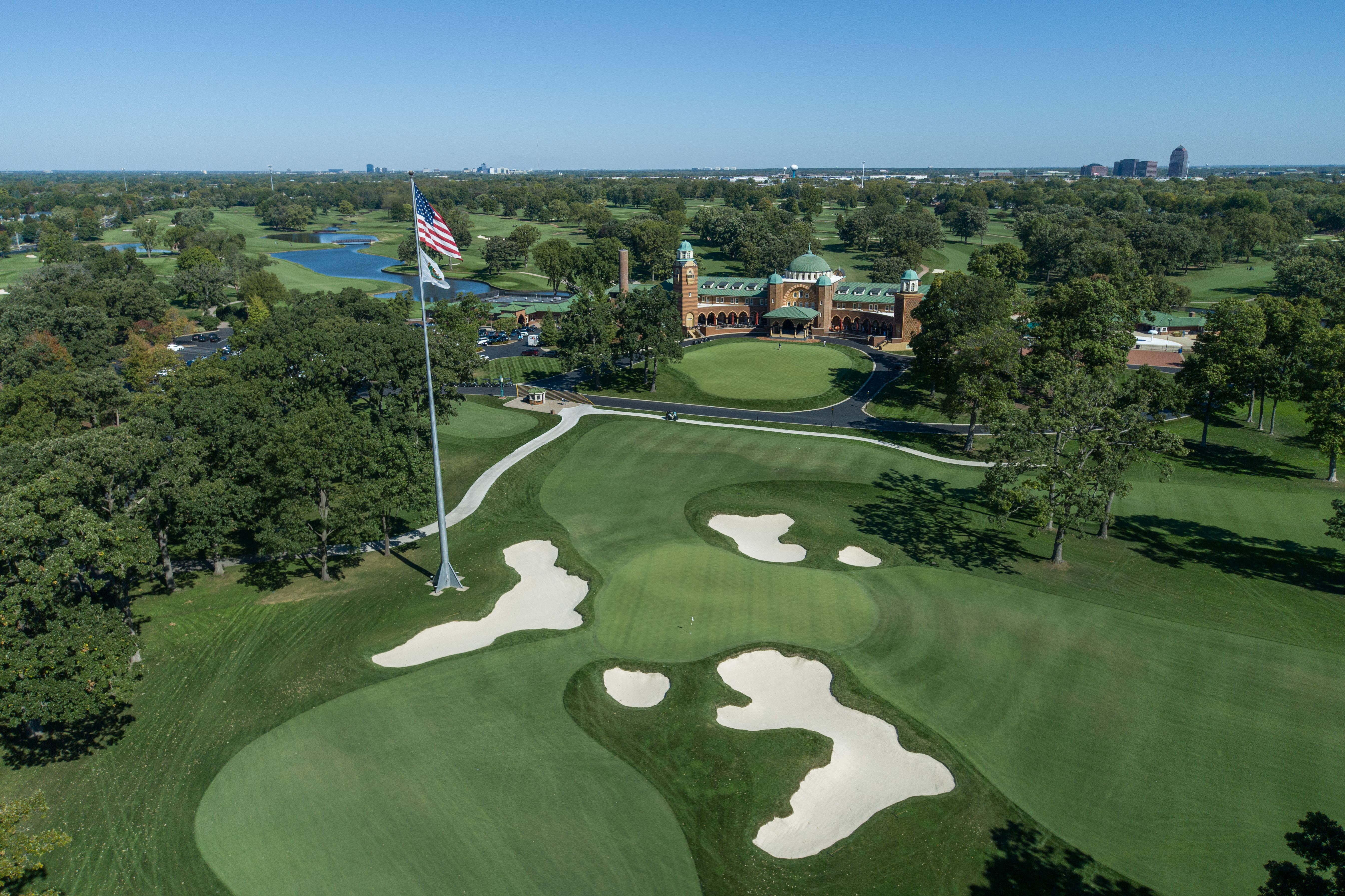 Aerial view of a pristine golf course with green fairways, bunkers, and a tall flagpole.