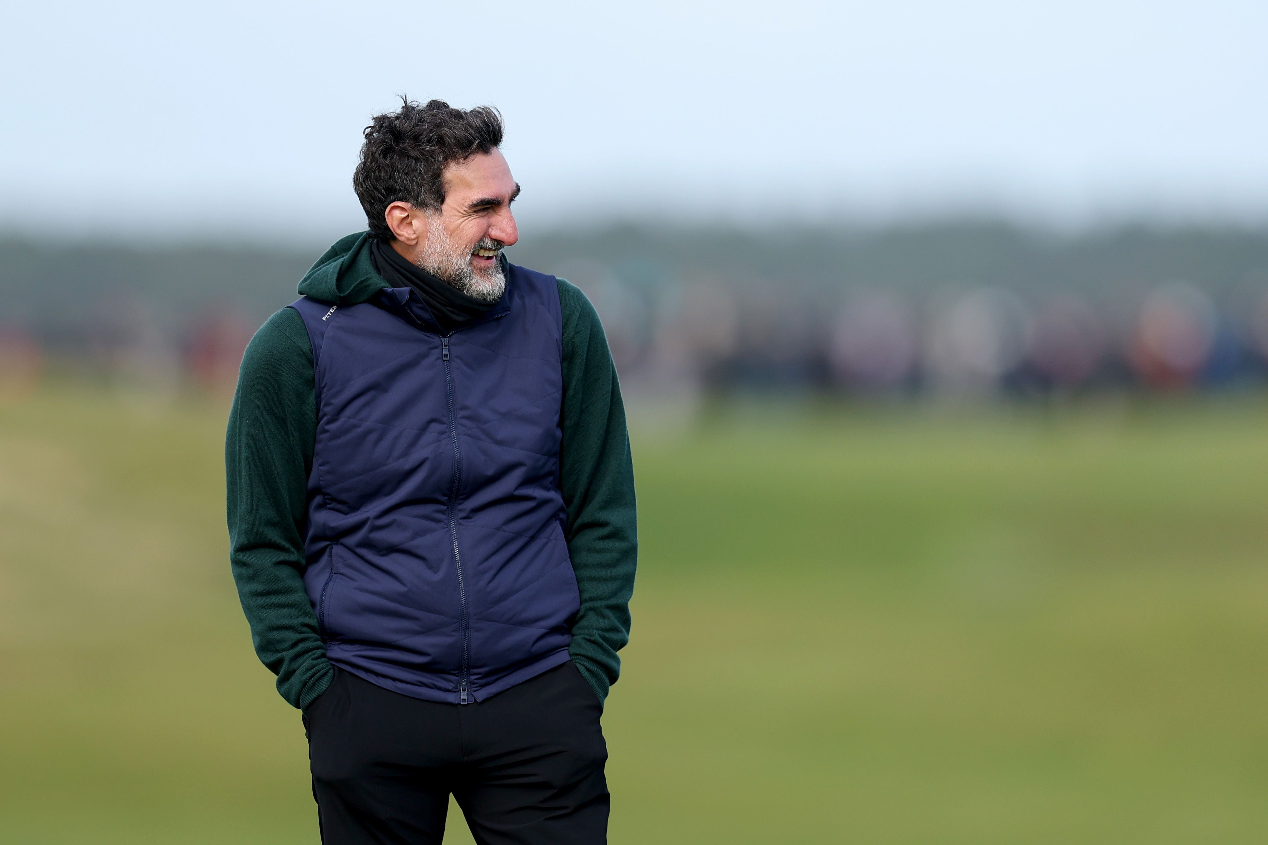 Man with dark hair and beard, wearing a vest, looking intently at a golf course.
