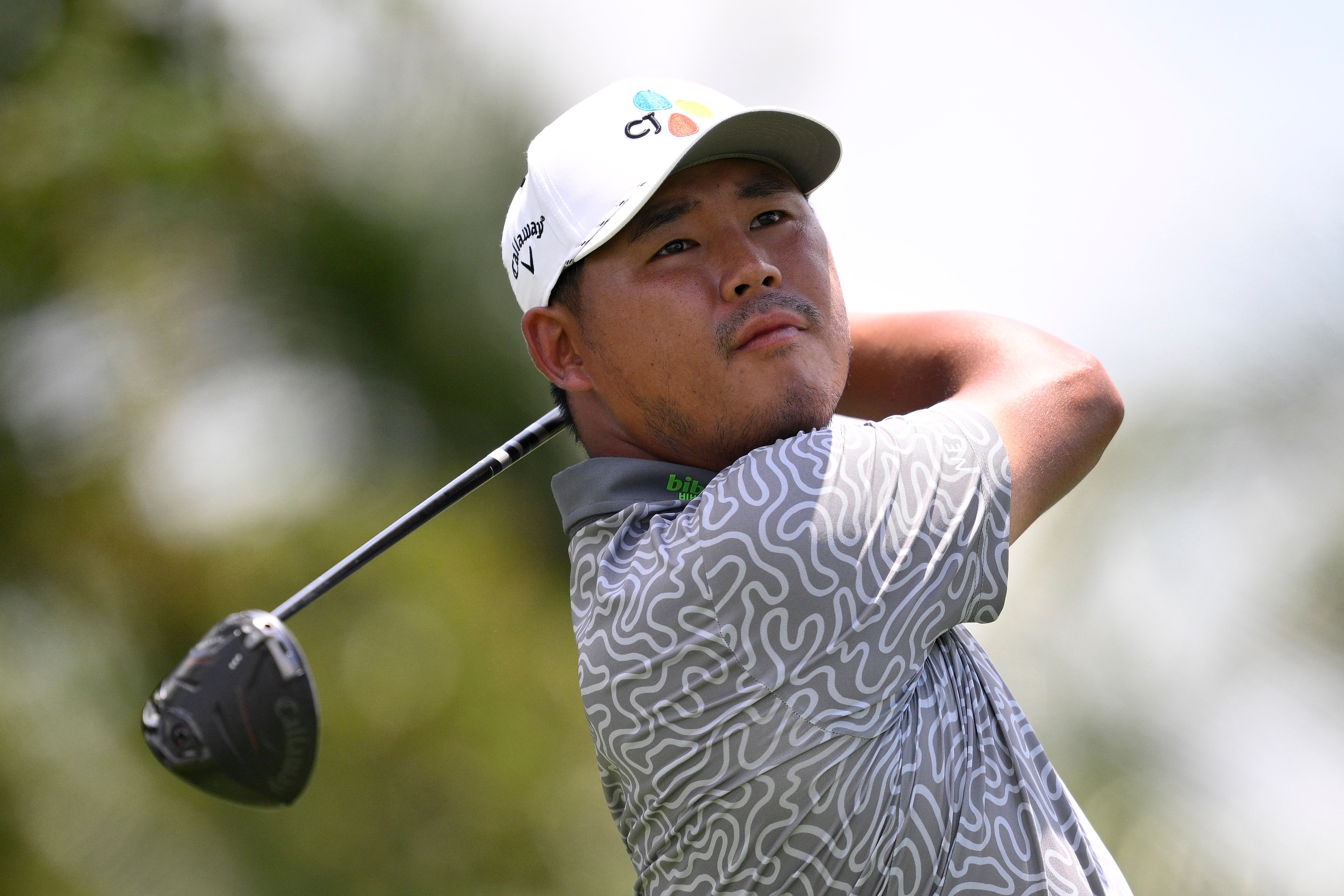 A male golfer in a white cap swings a driver, looking up at the ball's flight.
