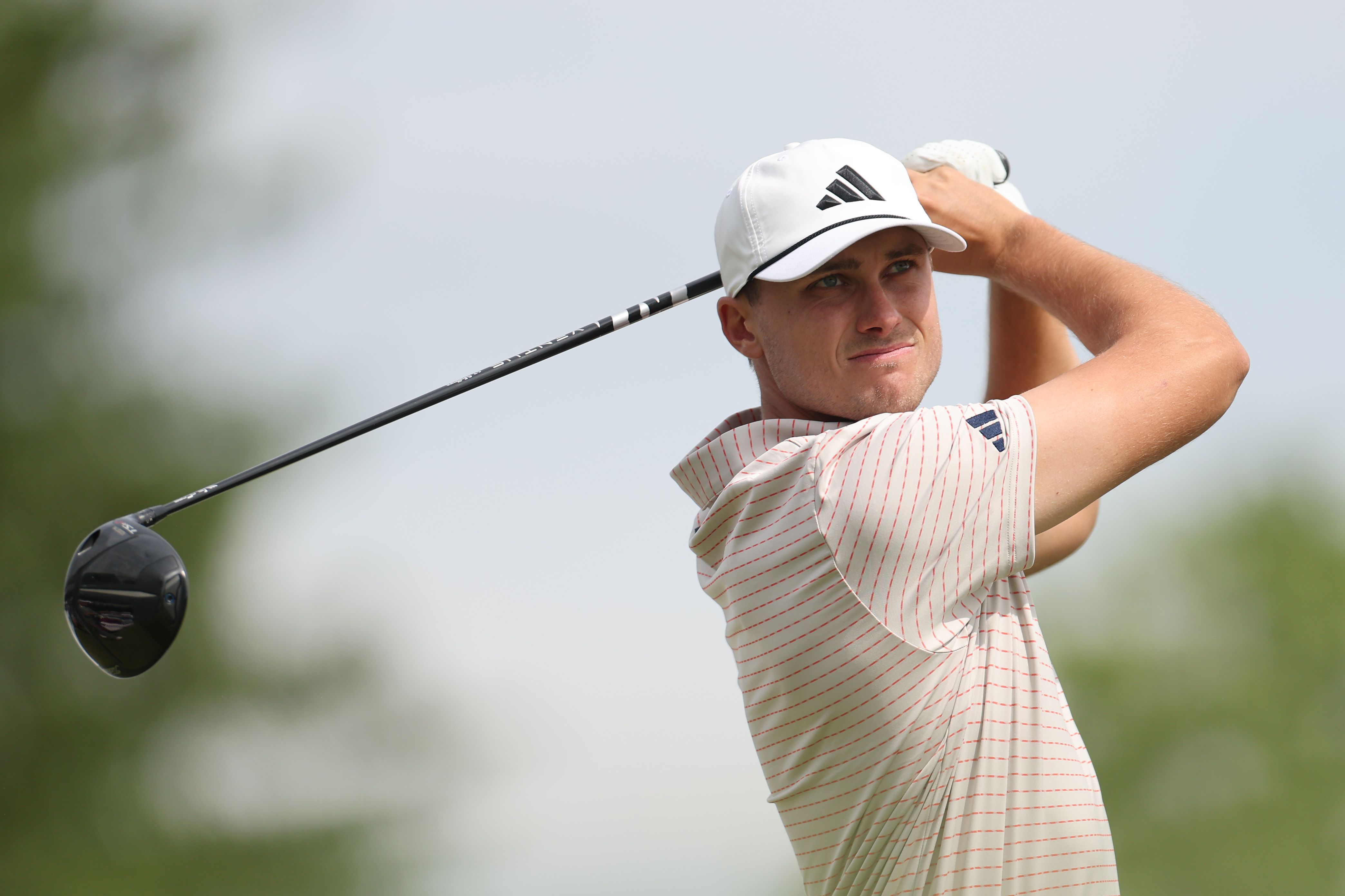 Male golfer in a white cap swings a driver on a bright golf course.