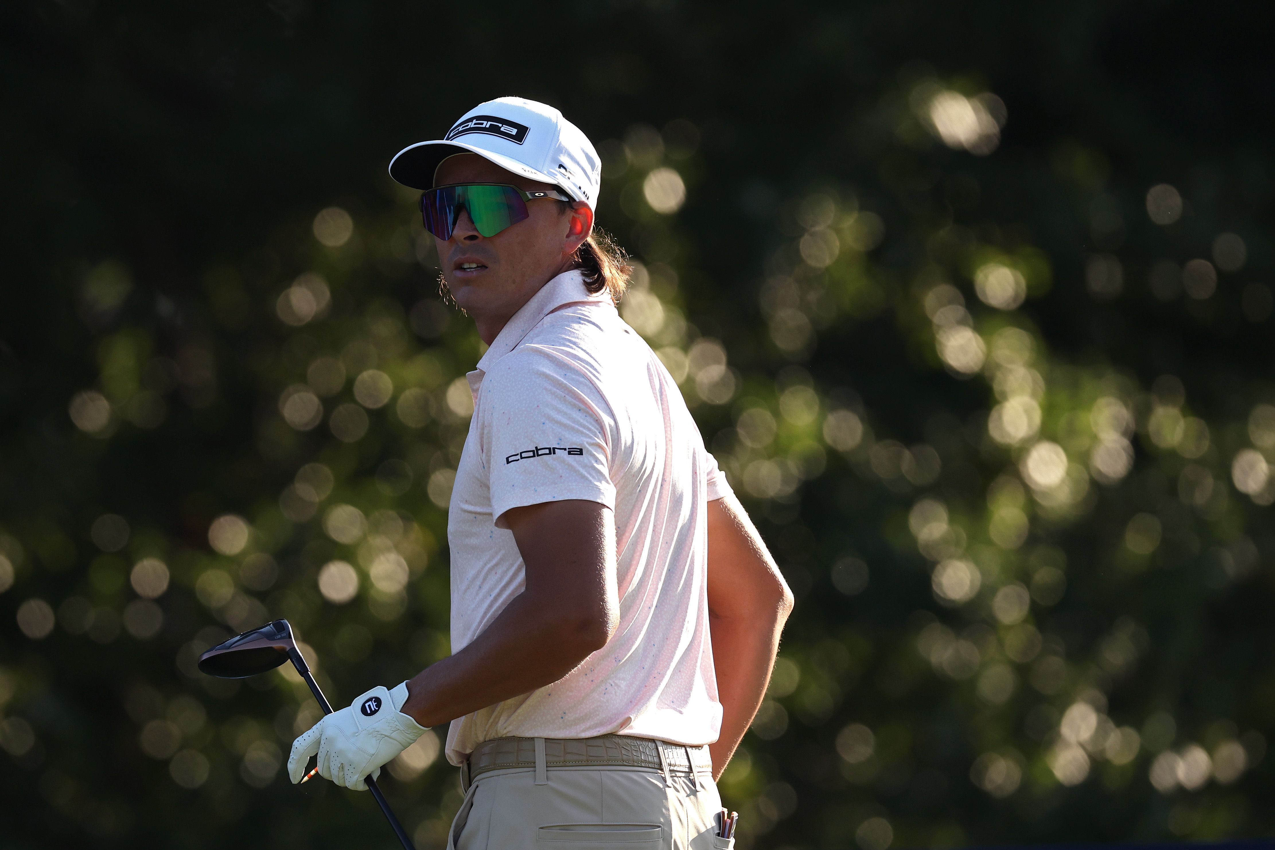 Male golfer in white shirt, cap, and sunglasses looks back on the course.