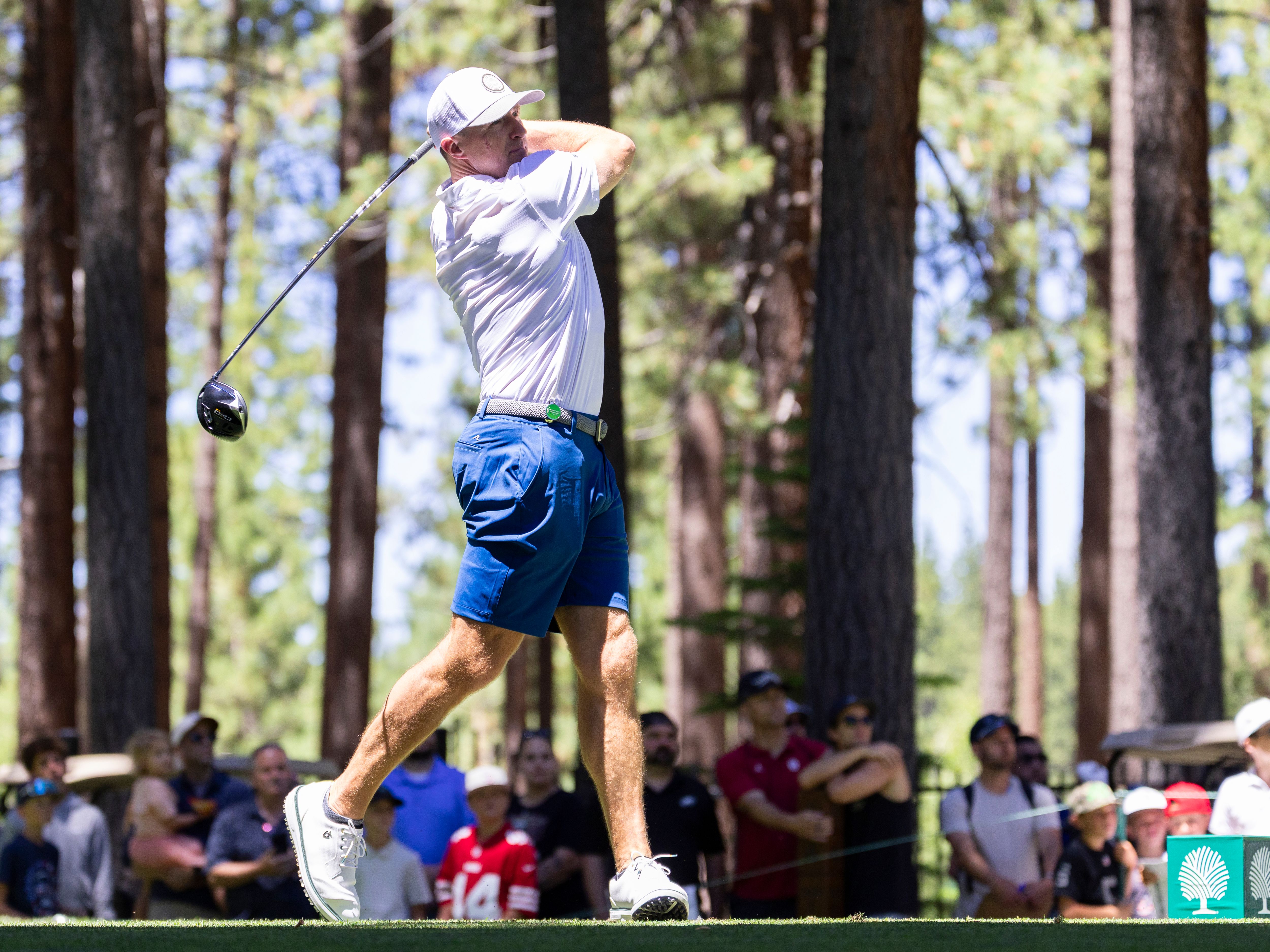 A male golfer in blue shorts and white shirt completes a powerful golf swing with a driver.