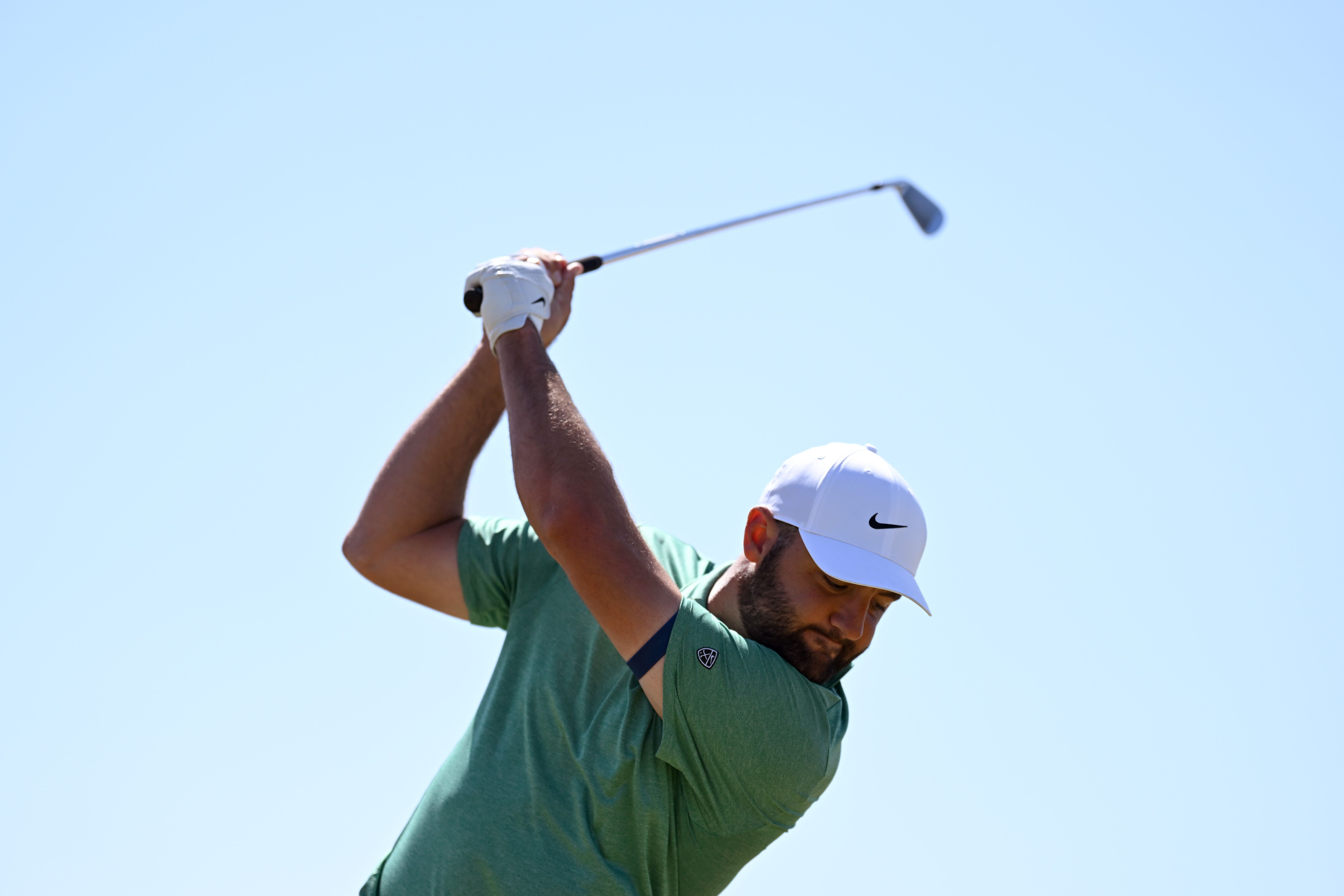 A male golfer in a green shirt and white cap swings a golf club against a clear blue sky.