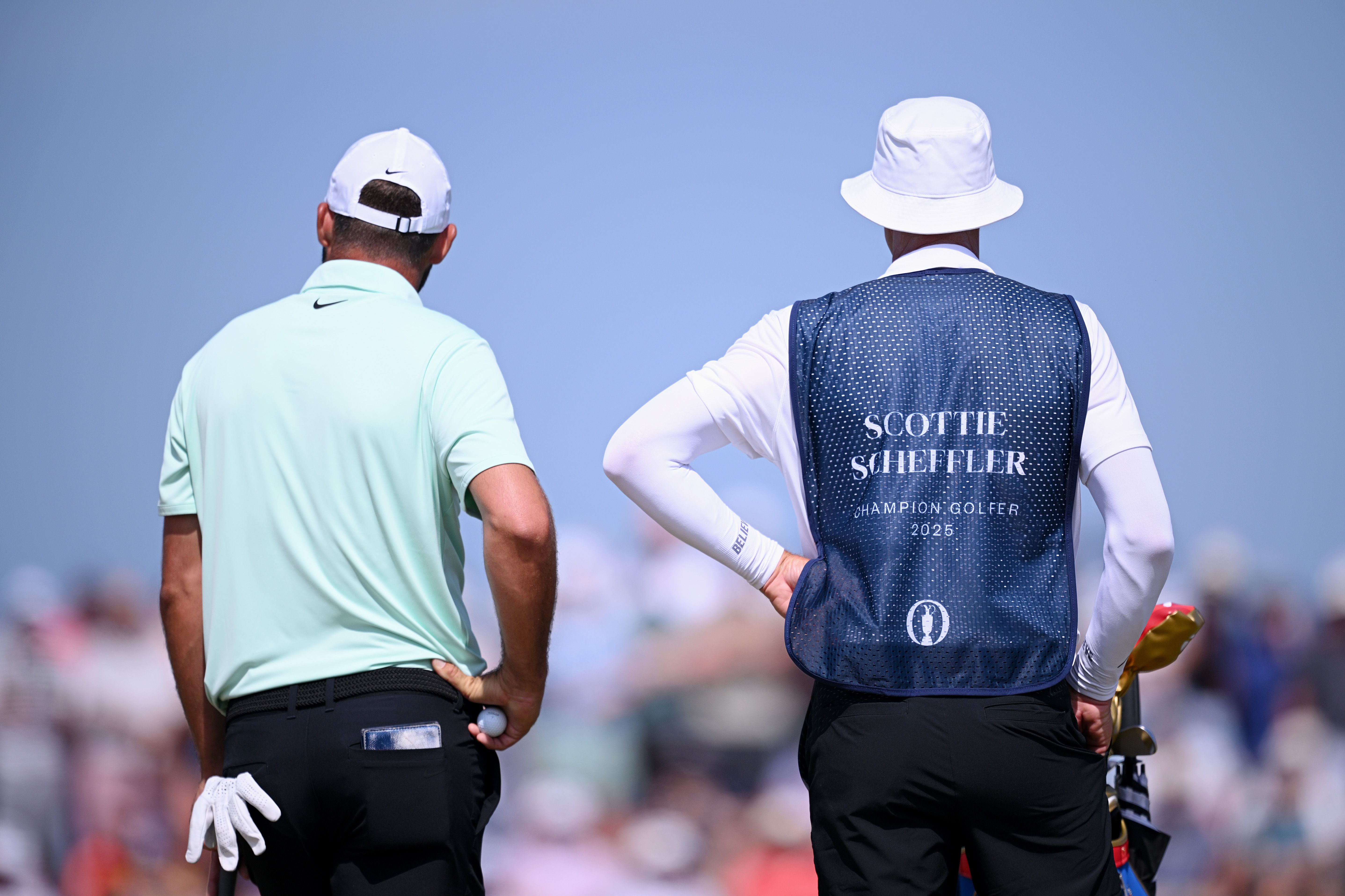 Two golfers, one wearing a caddie bib, stand on a course under a clear sky.