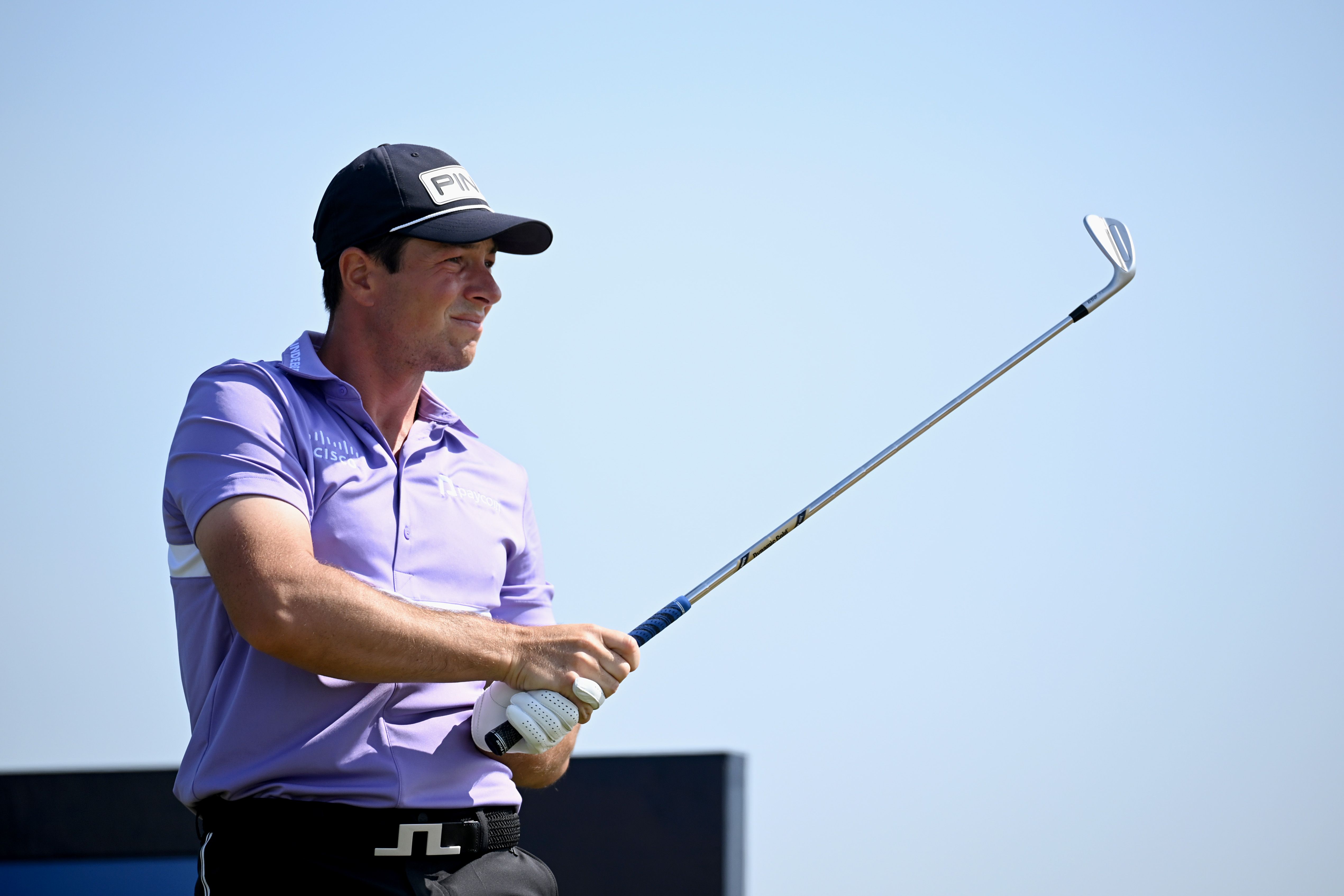 Male golfer in purple shirt and black cap holding a golf club under a clear sky.