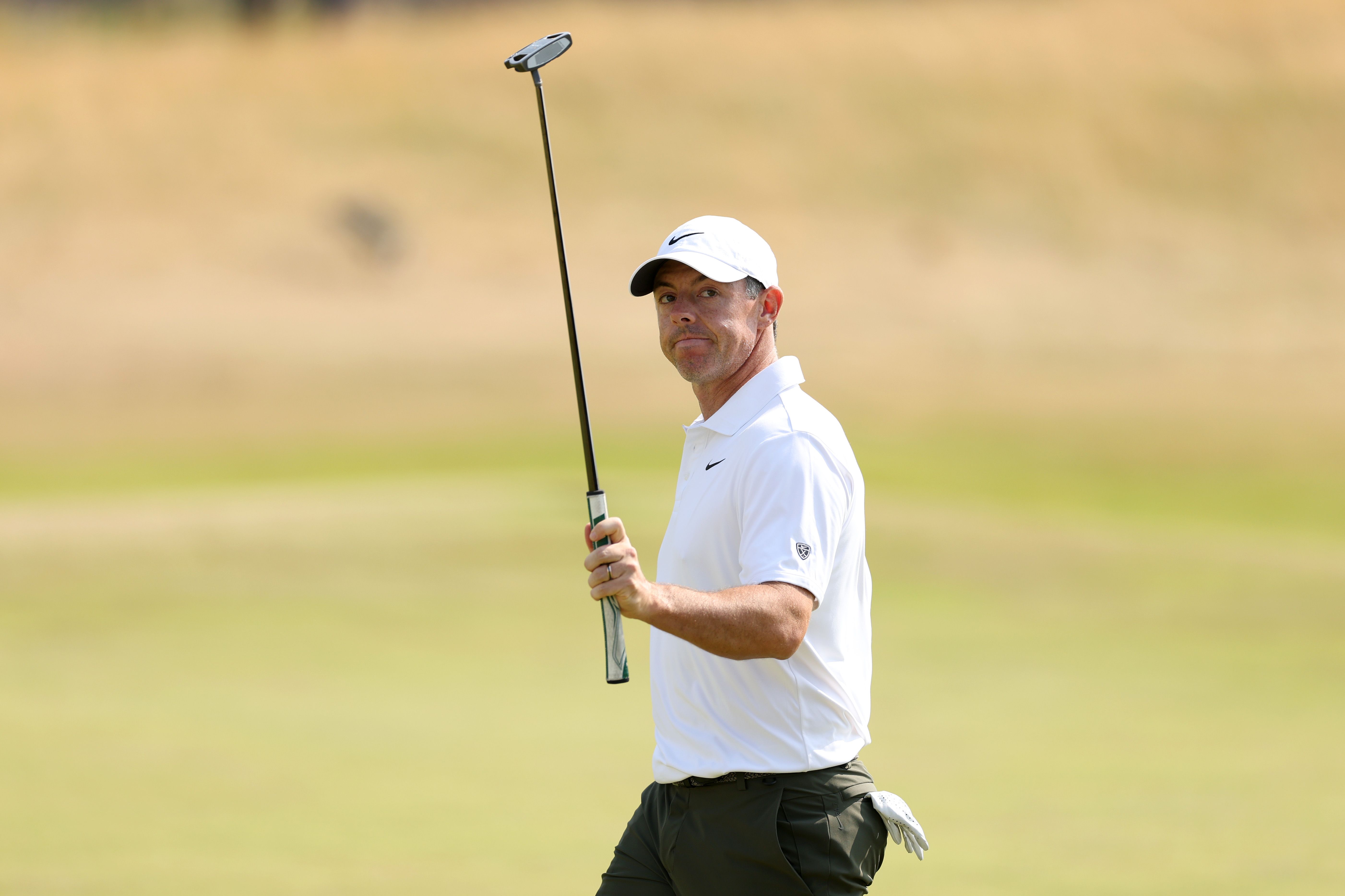 Male golfer in white shirt and cap walks on a green course, holding a putter.