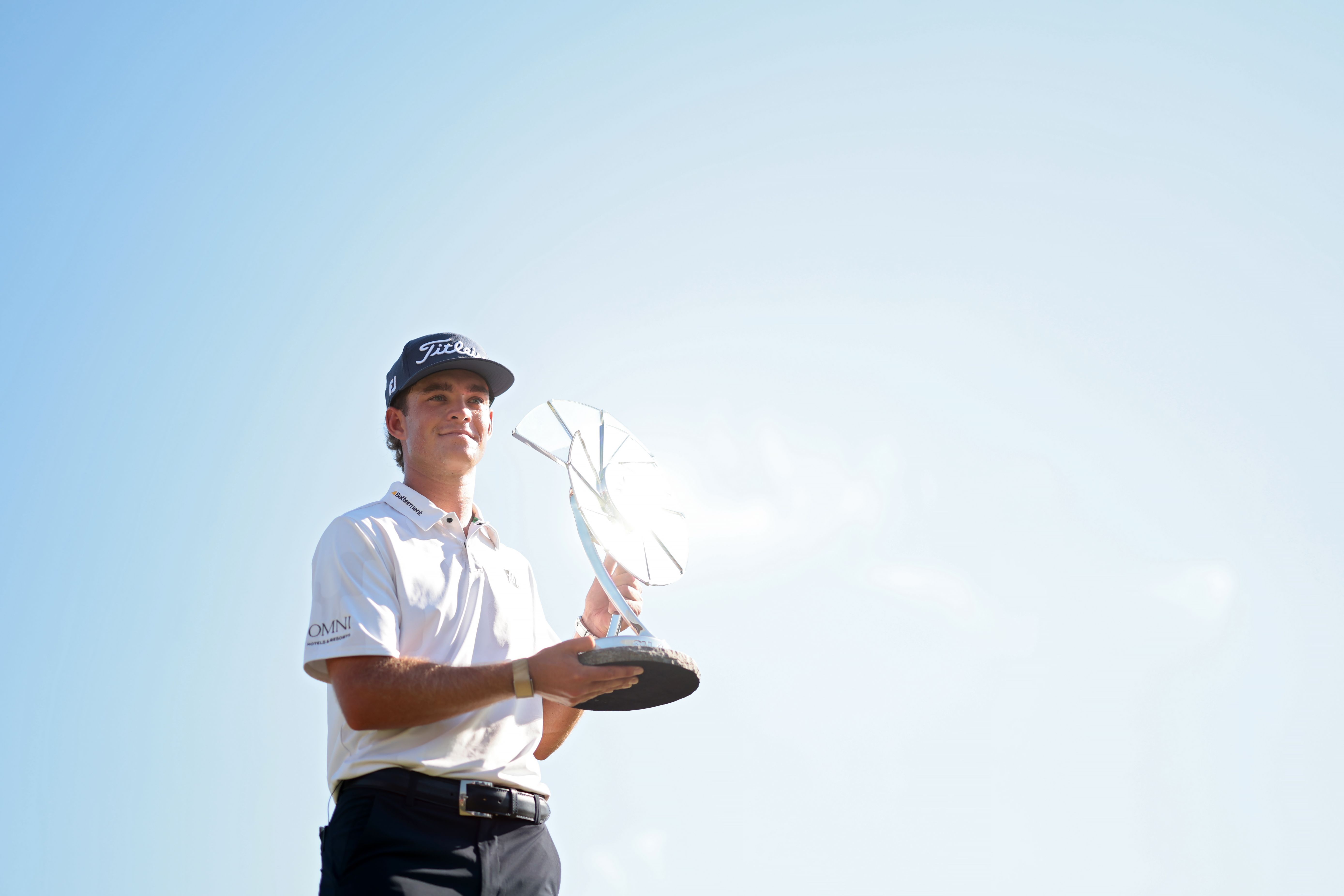 Man in a cap and polo shirt holding a trophy against a clear blue sky.