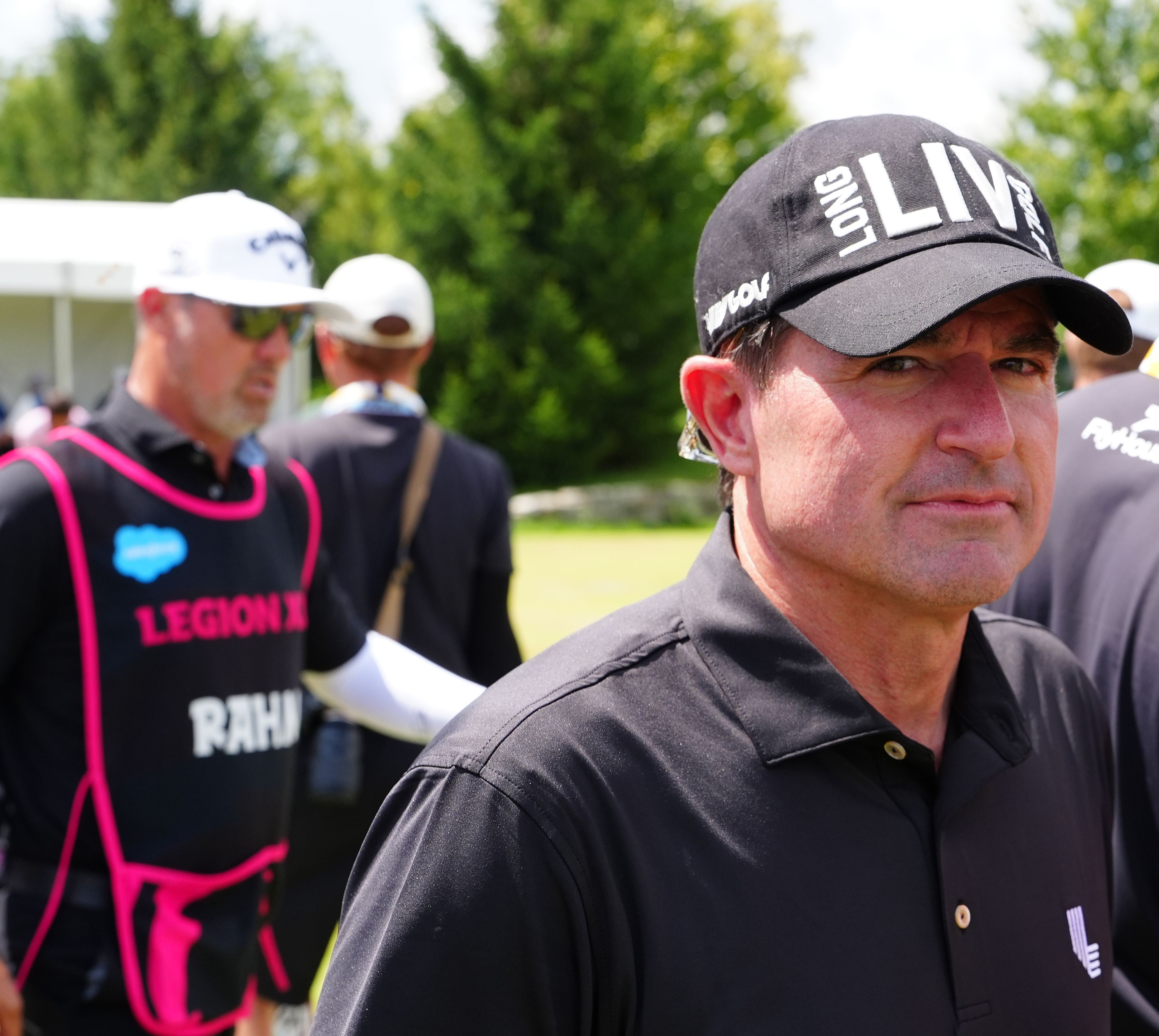 Man in black polo and LIV golf cap looks forward at an outdoor event.