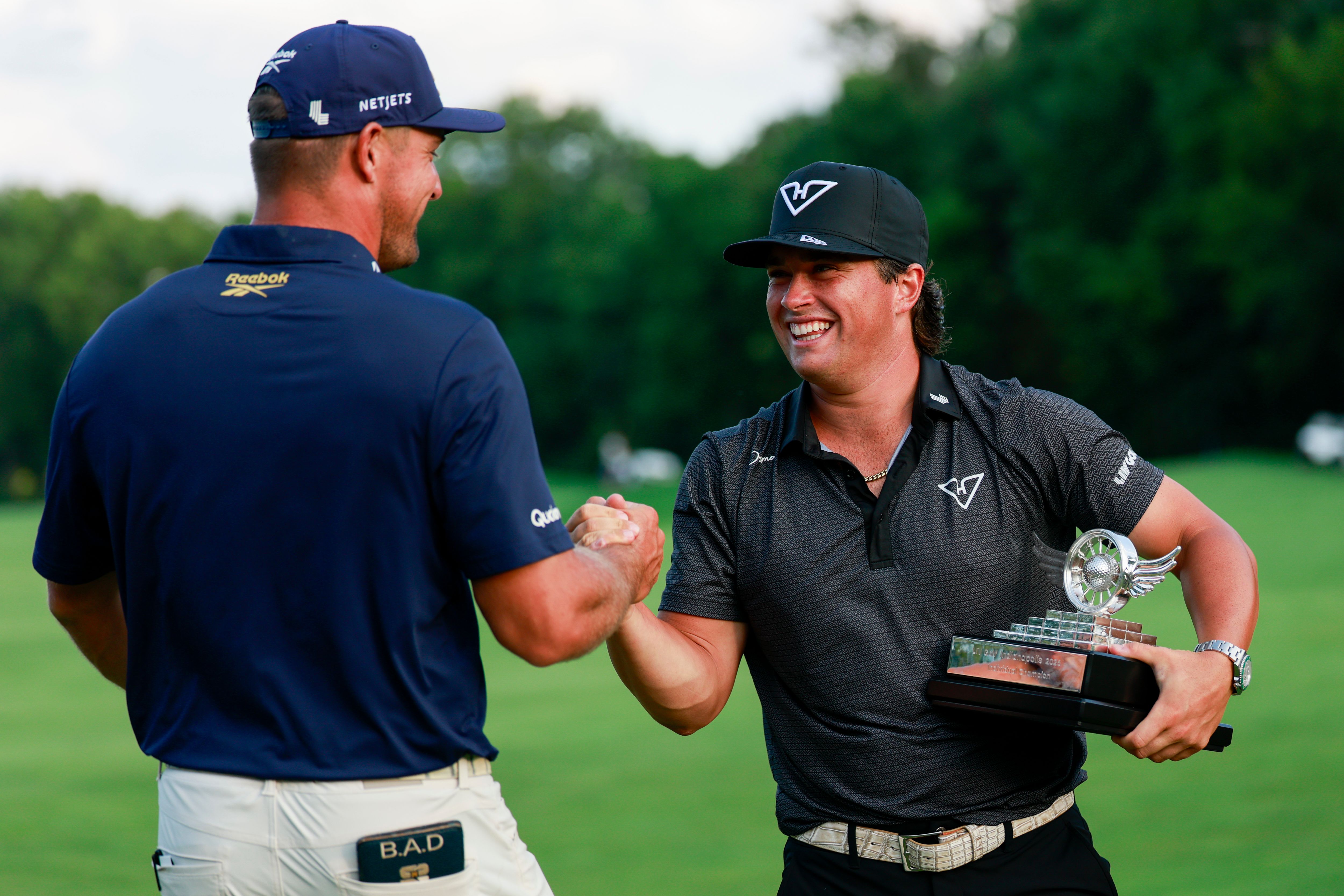 Two male golfers shake hands on a green, one holding a silver trophy after a match.