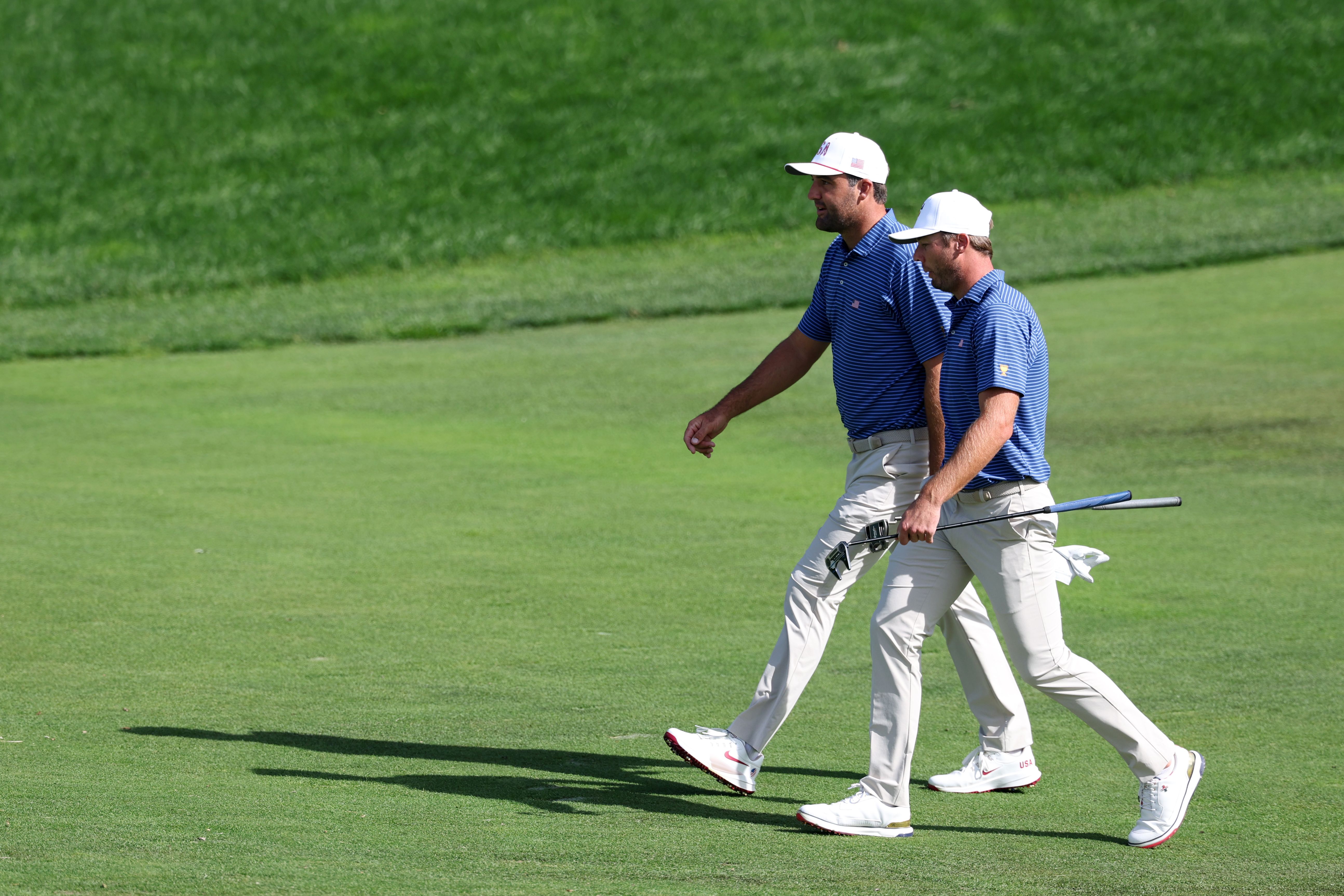 Two male golfers in blue shirts and white pants walk across a green golf course.