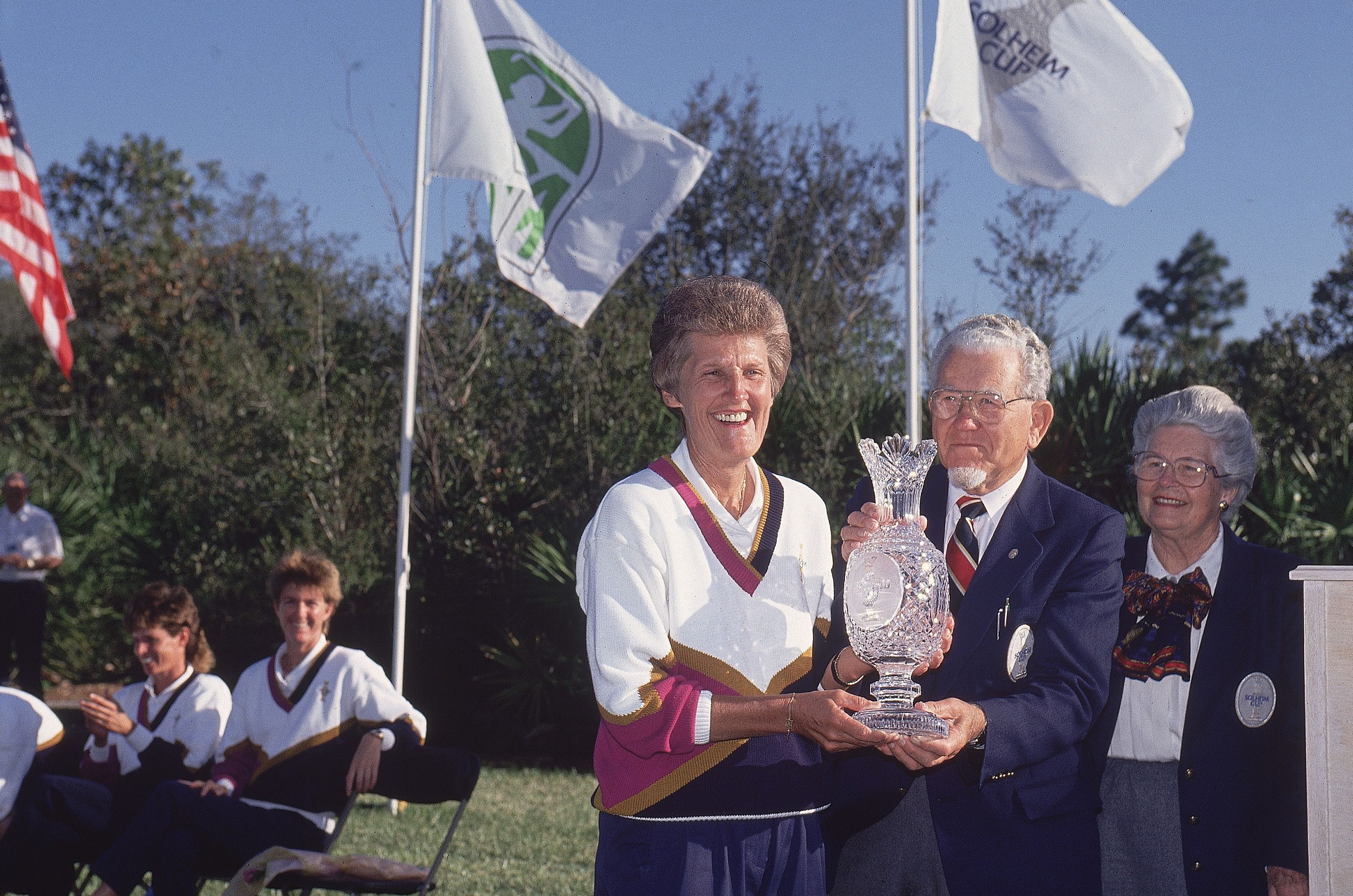 Woman in sports uniform receives large trophy from older man at outdoor ceremony.