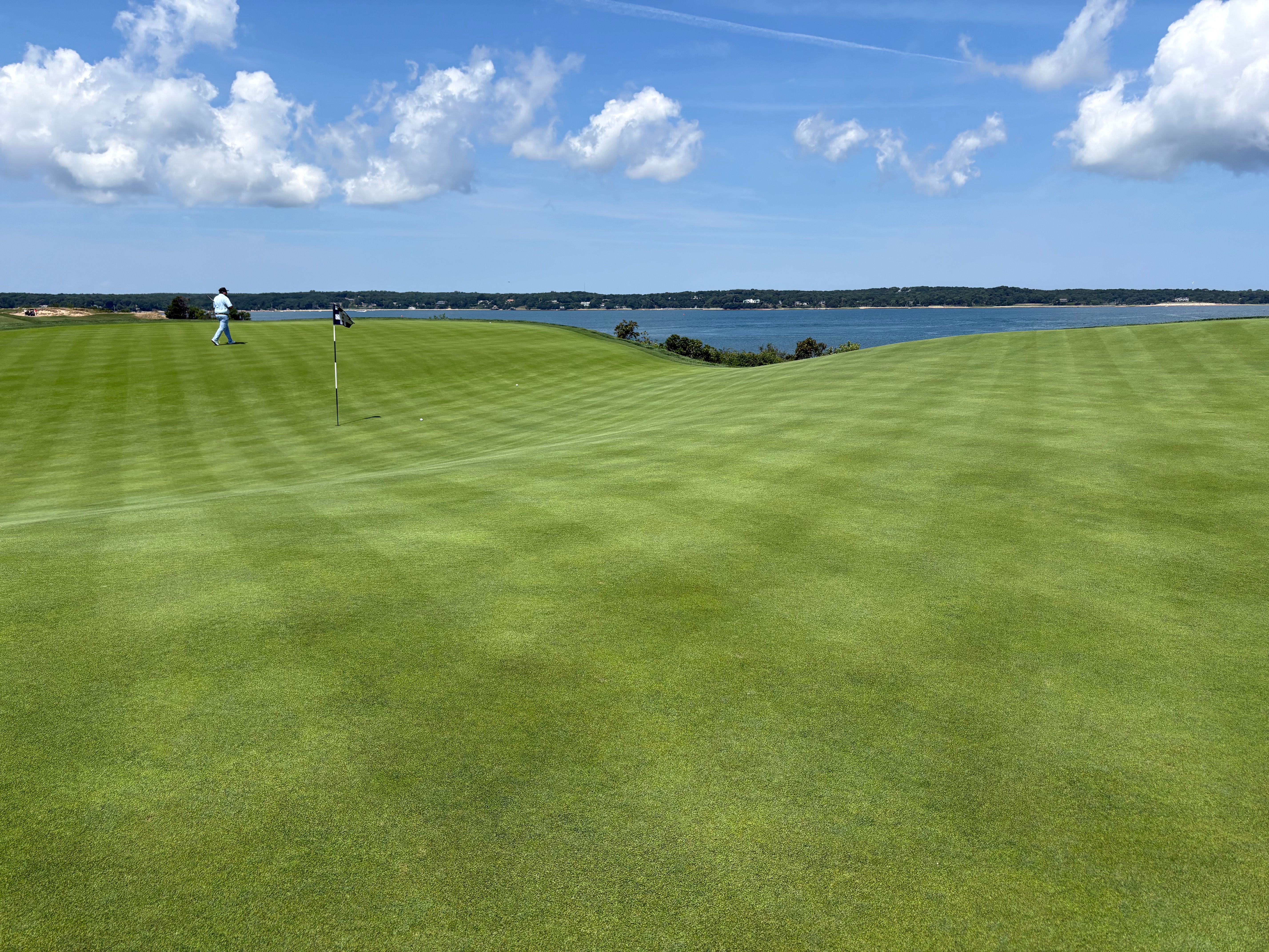 Vast green golf course with striped fairways overlooking a blue lake under a clear sky.