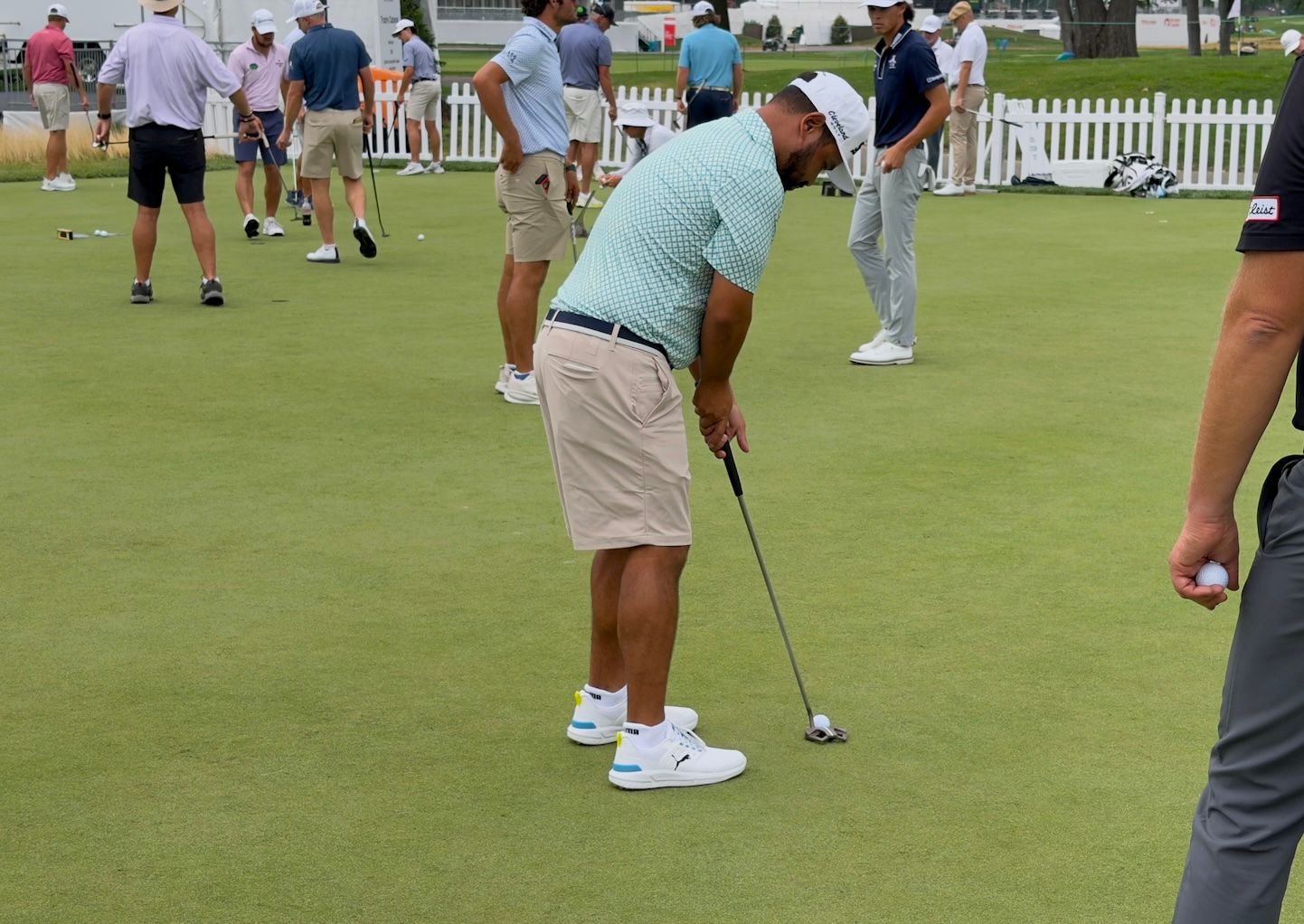 Golfer in light blue shirt and shorts putts on a green golf course with others nearby.