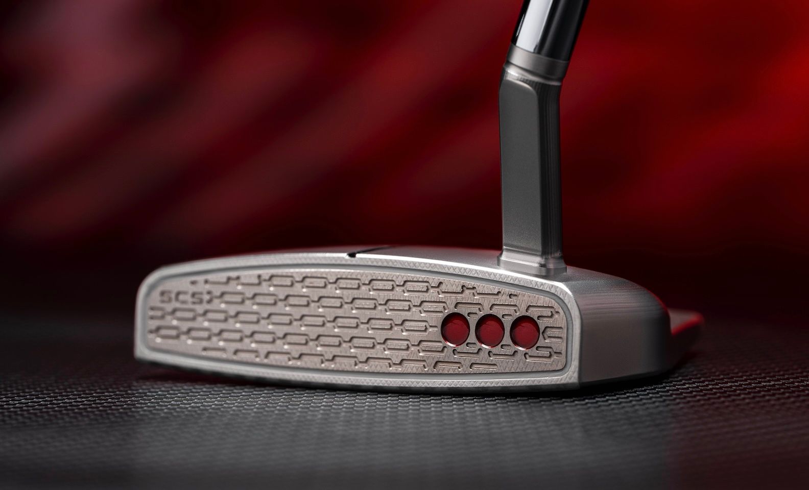 Silver golf putter head showing textured face and three red circular weights.