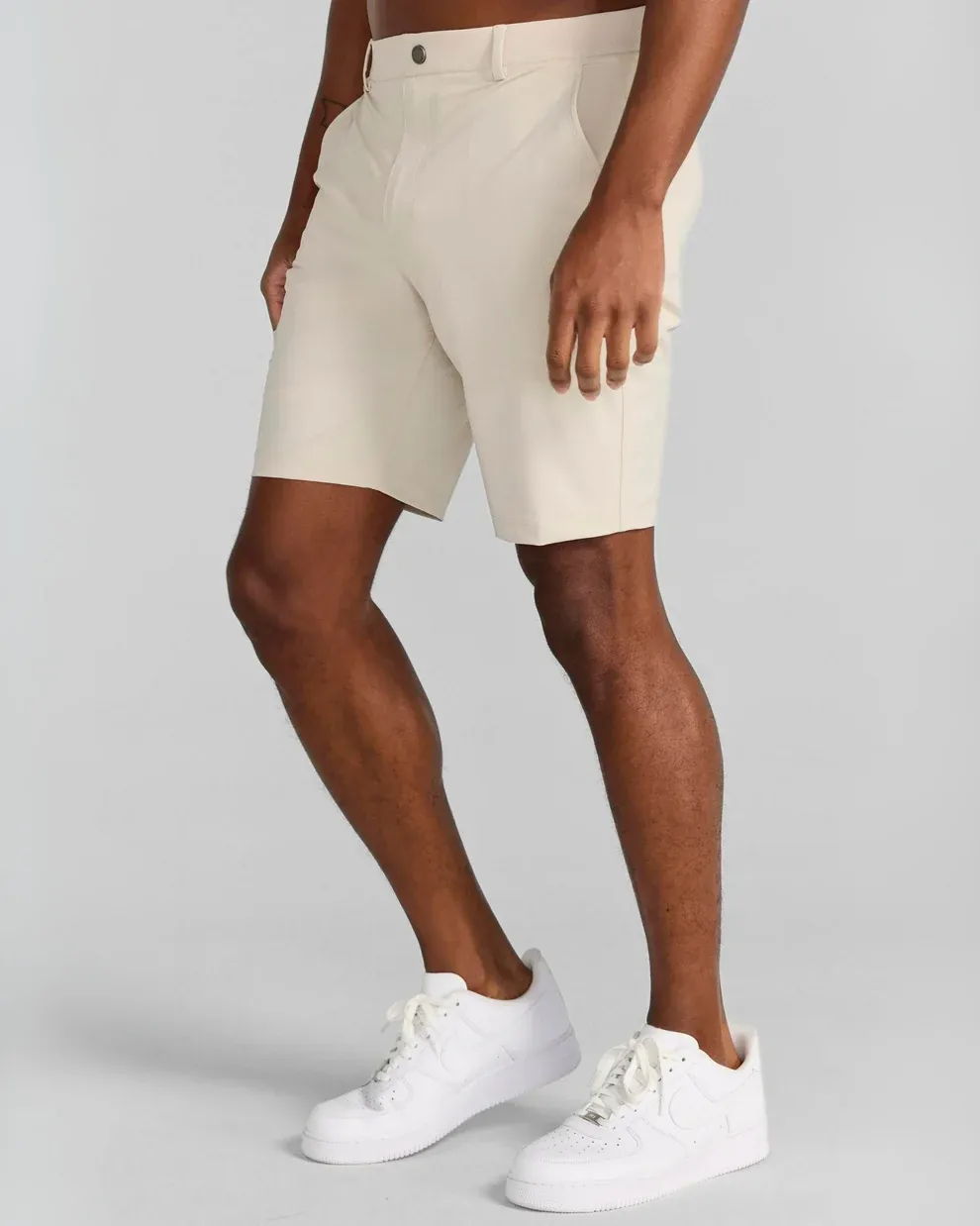 A person with dark skin wears beige shorts and white sneakers, standing against a light gray background.