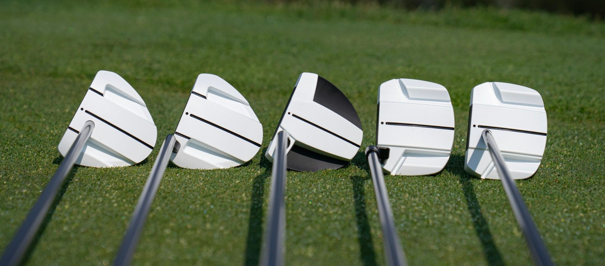 Five white golf putters with black alignment lines lined up on green grass.