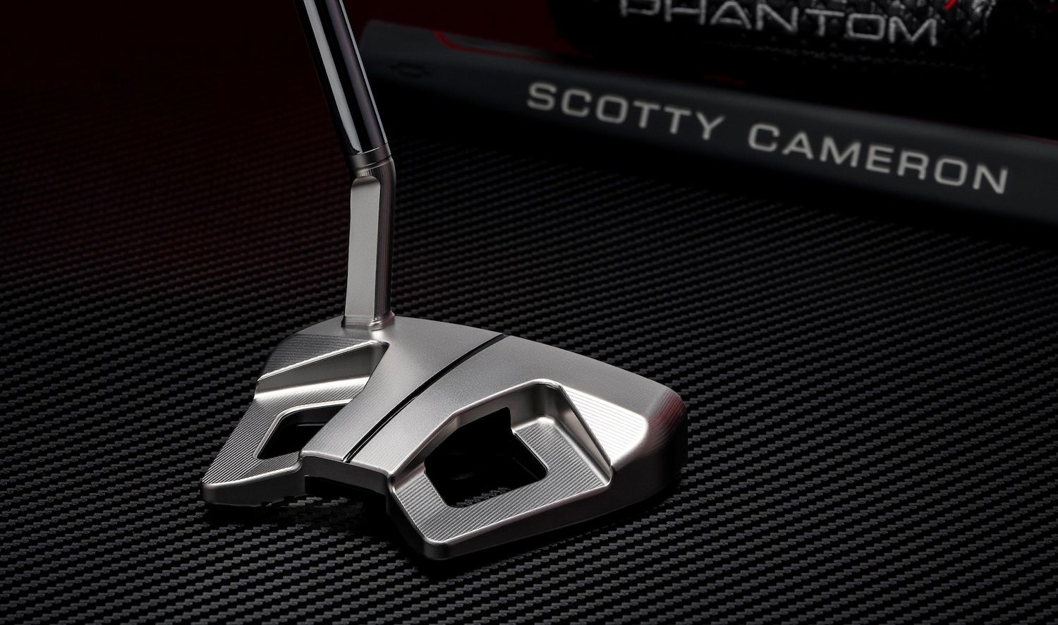 Close-up of a modern silver golf putter head with sharp angles on a dark surface.