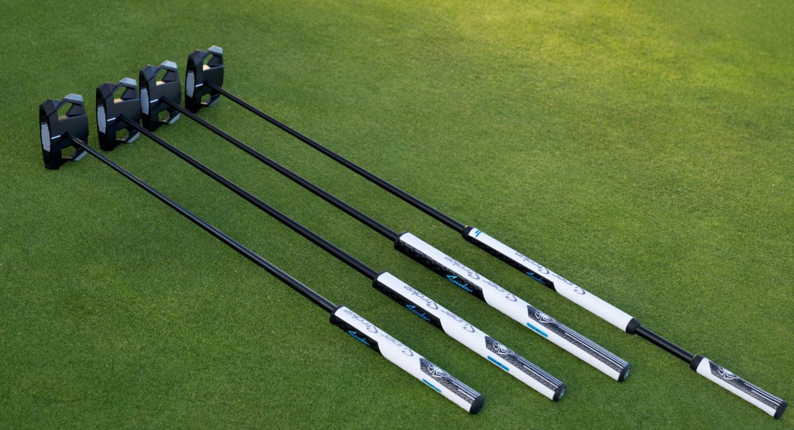Four modern golf putters with black heads and silver shafts resting on green grass.