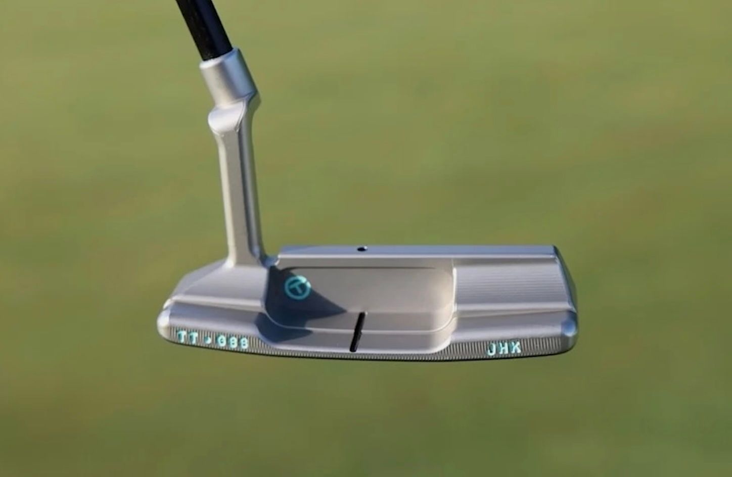 A silver golf putter head with a black shaft rests on vibrant green grass.