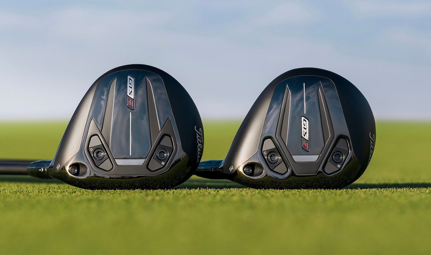 Two dark golf club driver heads resting side-by-side on green grass.