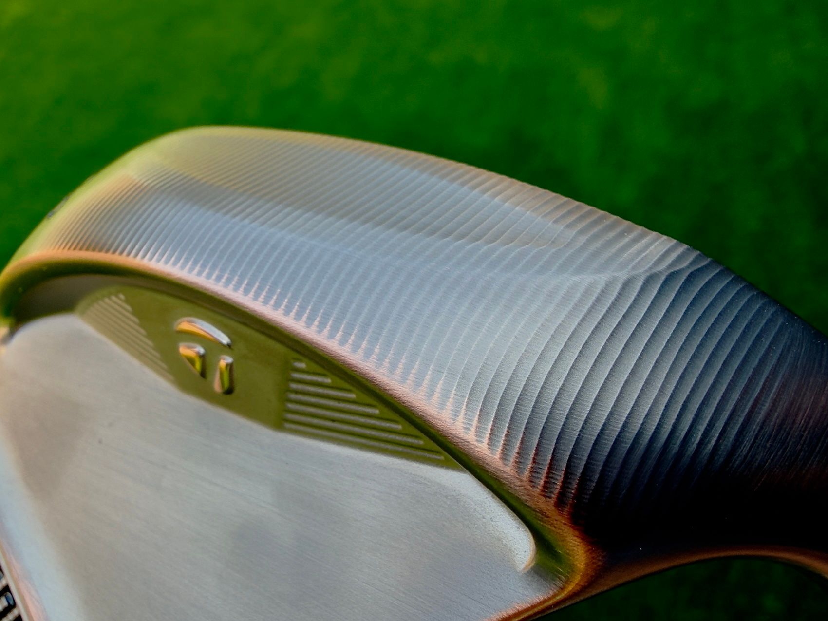A metallic TaylorMade golf club head features textured grooves on a green backdrop.