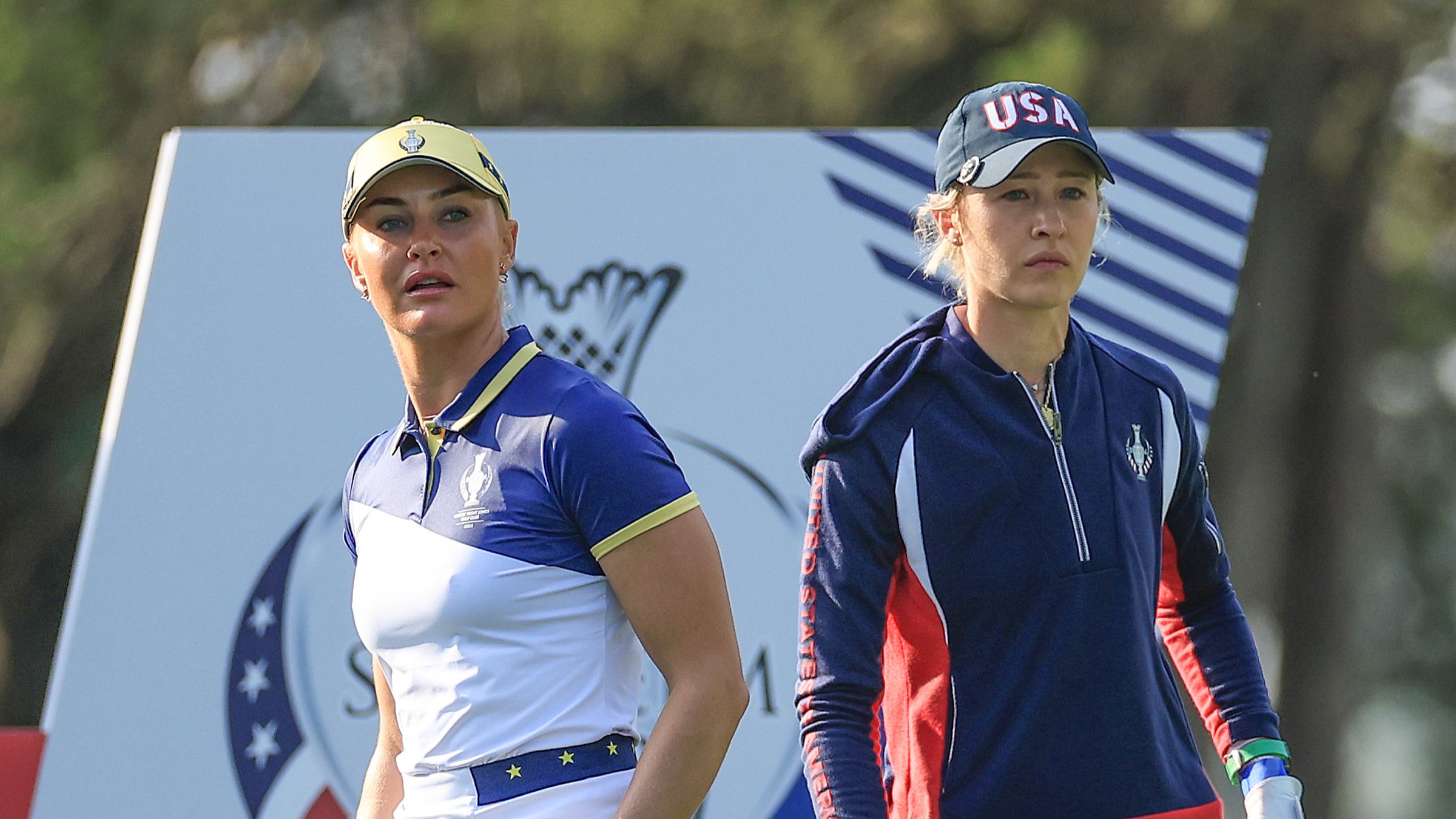 Two female golfers, wearing athletic attire, stand intently on a sunny golf course.